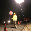 Traffic management worker in reflective vest holding a stop-go sign, with a Powermoon lighting unit positioned nearby on the road